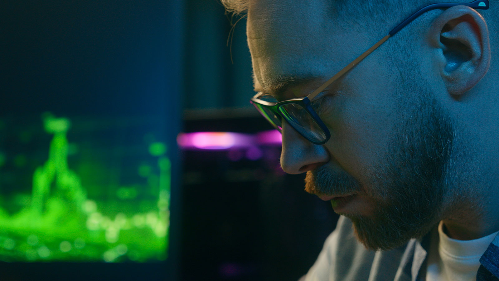 Close-up profile of a man wearing glasses, looking down in concentration, with a glowing green data graph behind him