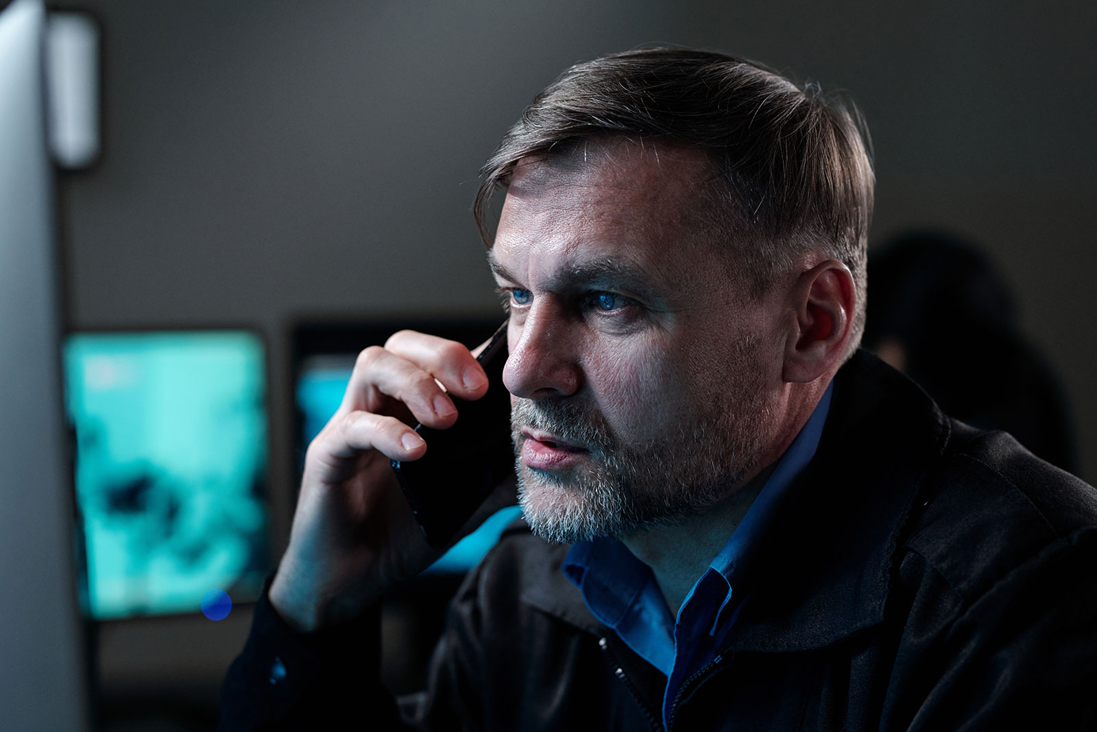 Man on a phone call looking intently at a screen, with monitors glowing in the dimly lit room behind him