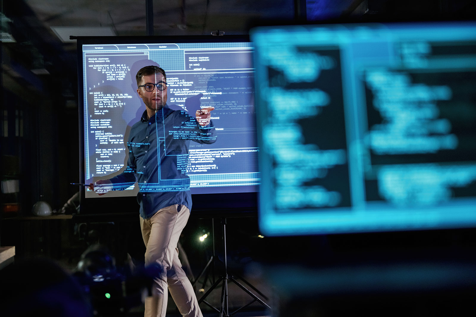 Man presenting in front of a projection screen of code, pointing towards the audience as the code is projected across him