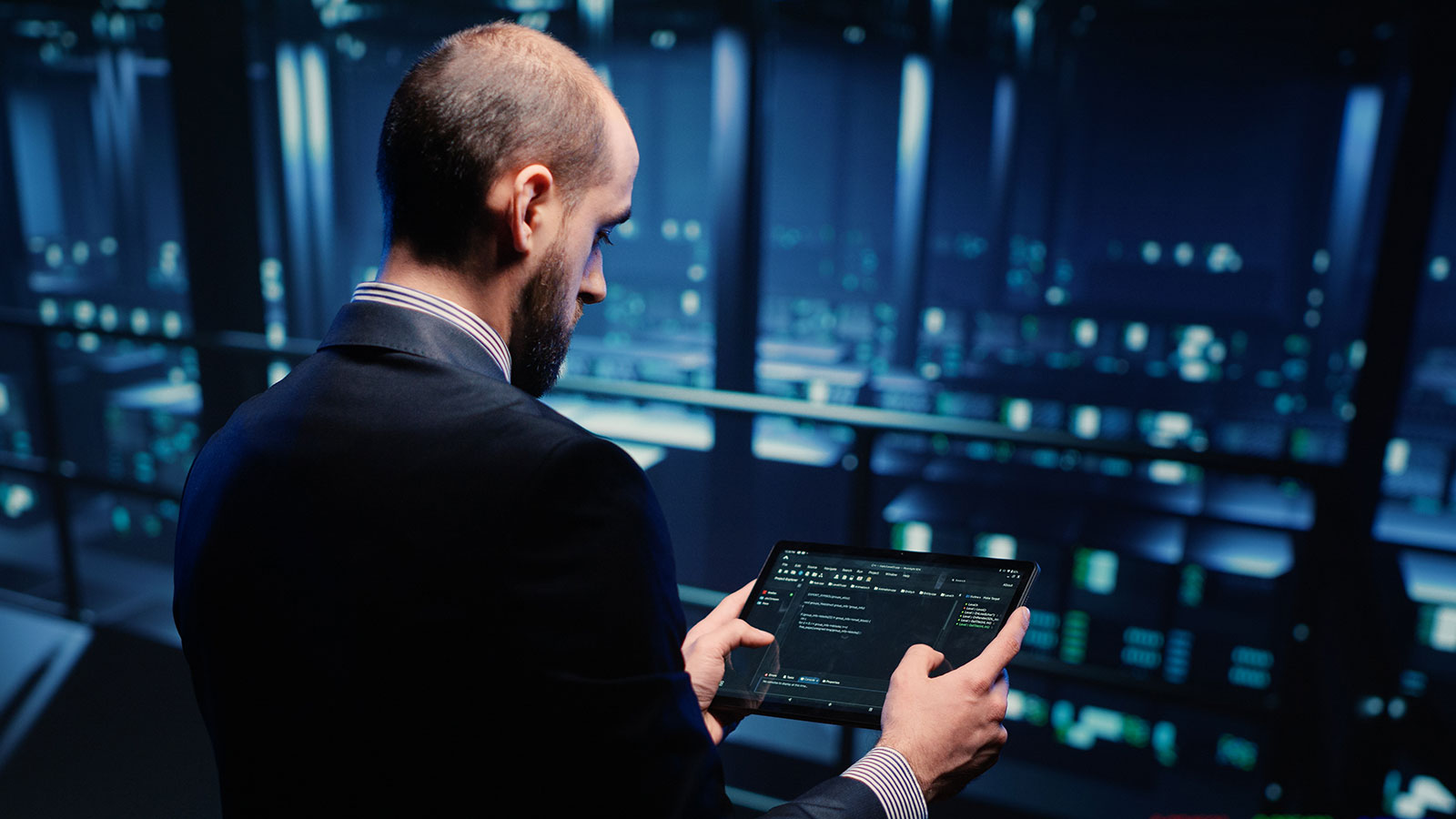 Man in a suit checking code on a tablet as he stands in a data centre, surrounded by rows of illuminated servers