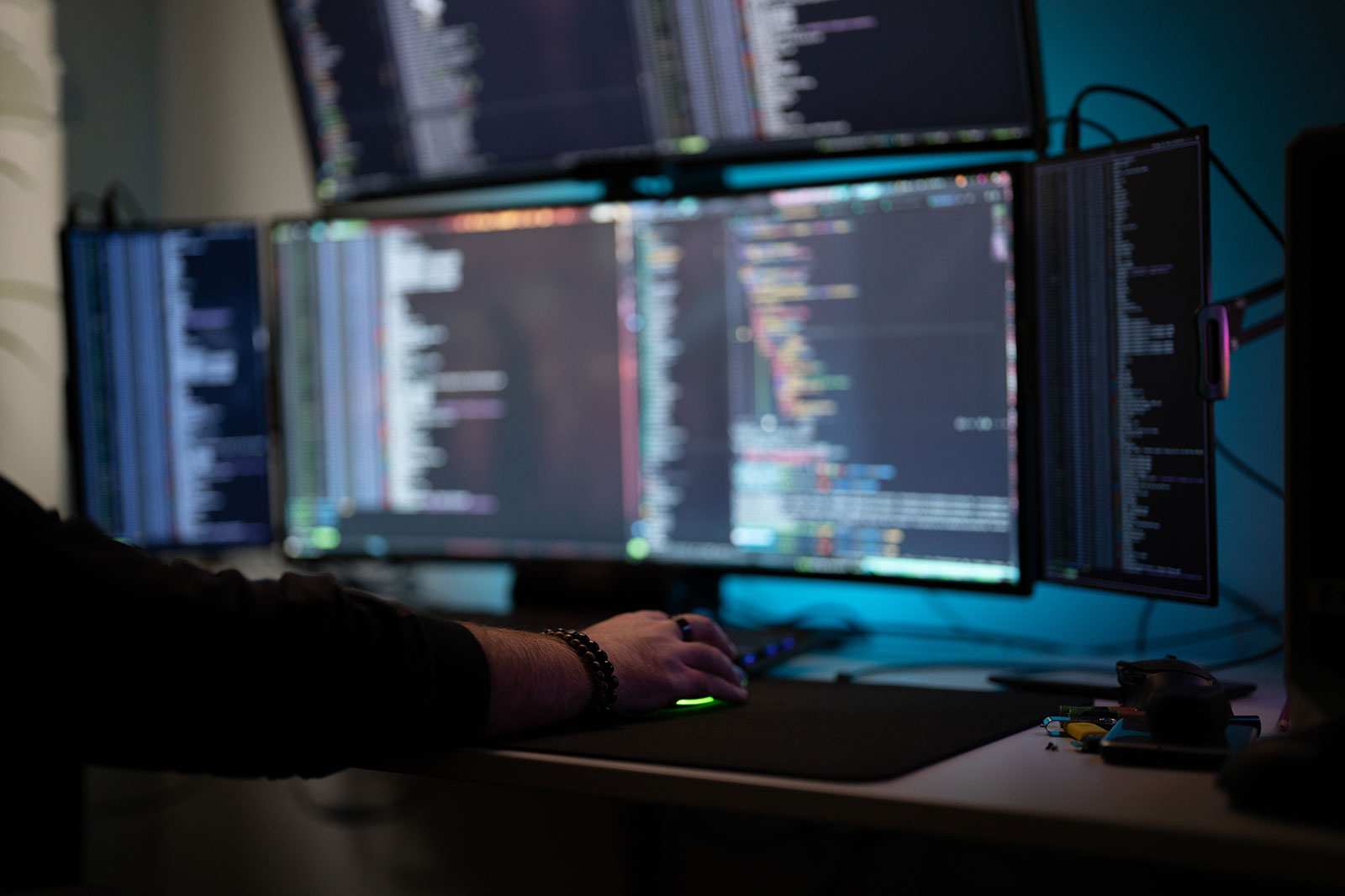 Hand on a glowing mouse at a desk with four monitors, all displaying code and terminal output