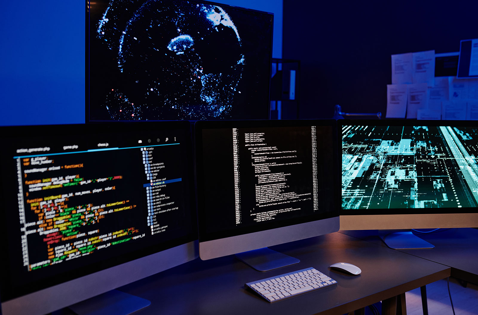Dimly lit workstation with three monitors showing source code and data visualisations, beneath a large screen of a digital globe