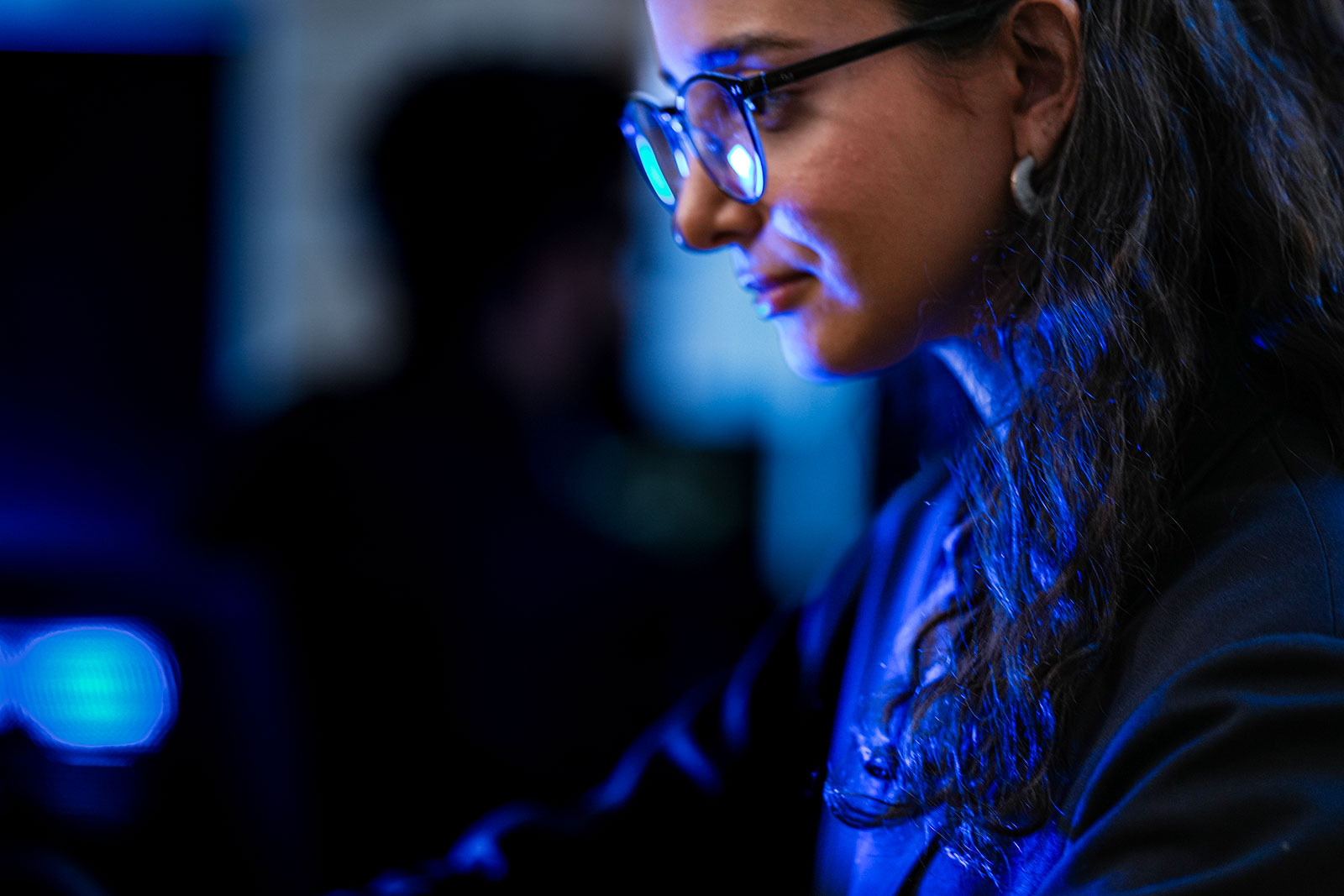 Woman wearing glasses, working intently at a screen, her face lit by blue light in a dimly lit room