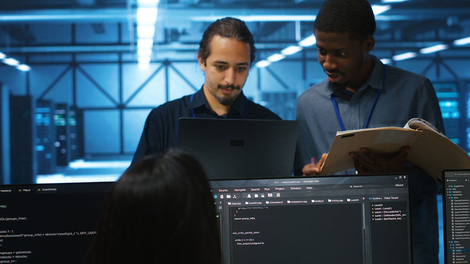Two engineers reviewing a laptop and a printed binder together, with code on monitors in the foreground and server racks behind