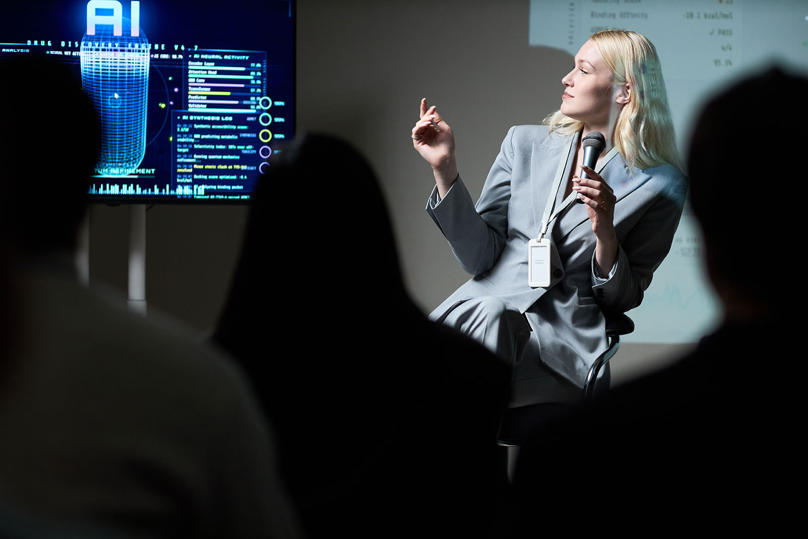 Woman with a microphone presenting to an audience, gesturing towards a screen showing an AI analytics dashboard