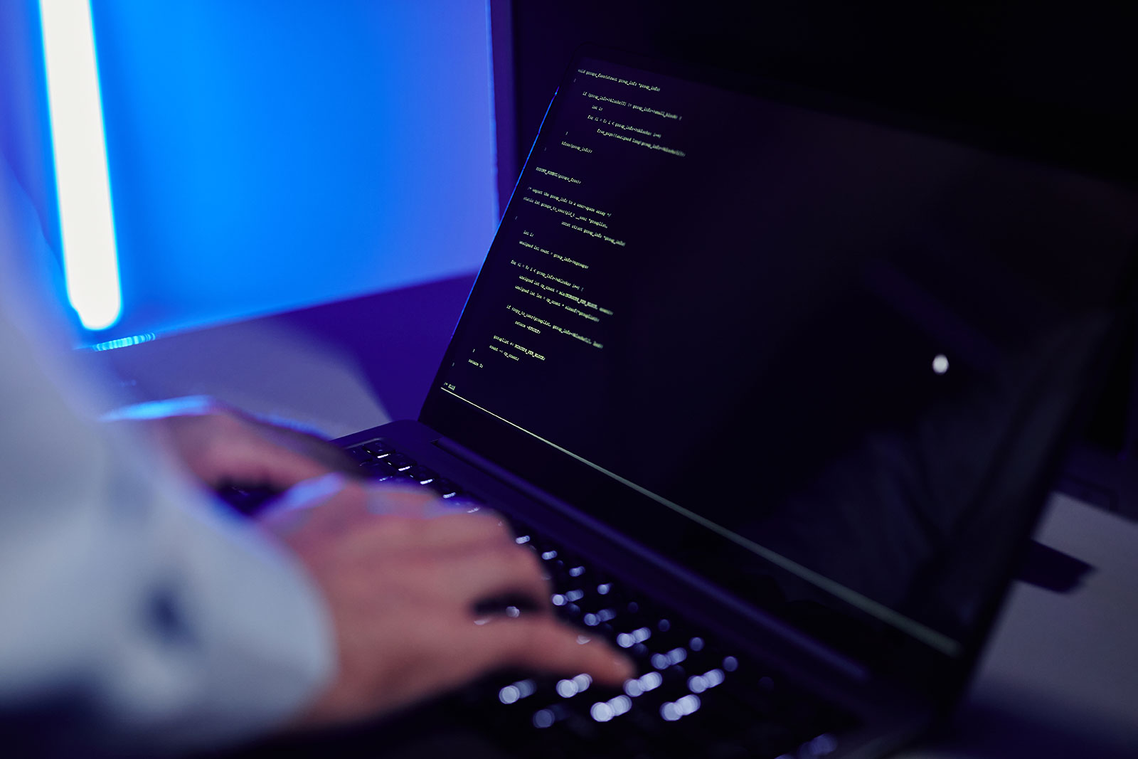 Close-up of hands typing on a laptop displaying lines of code, lit by blue neon light
