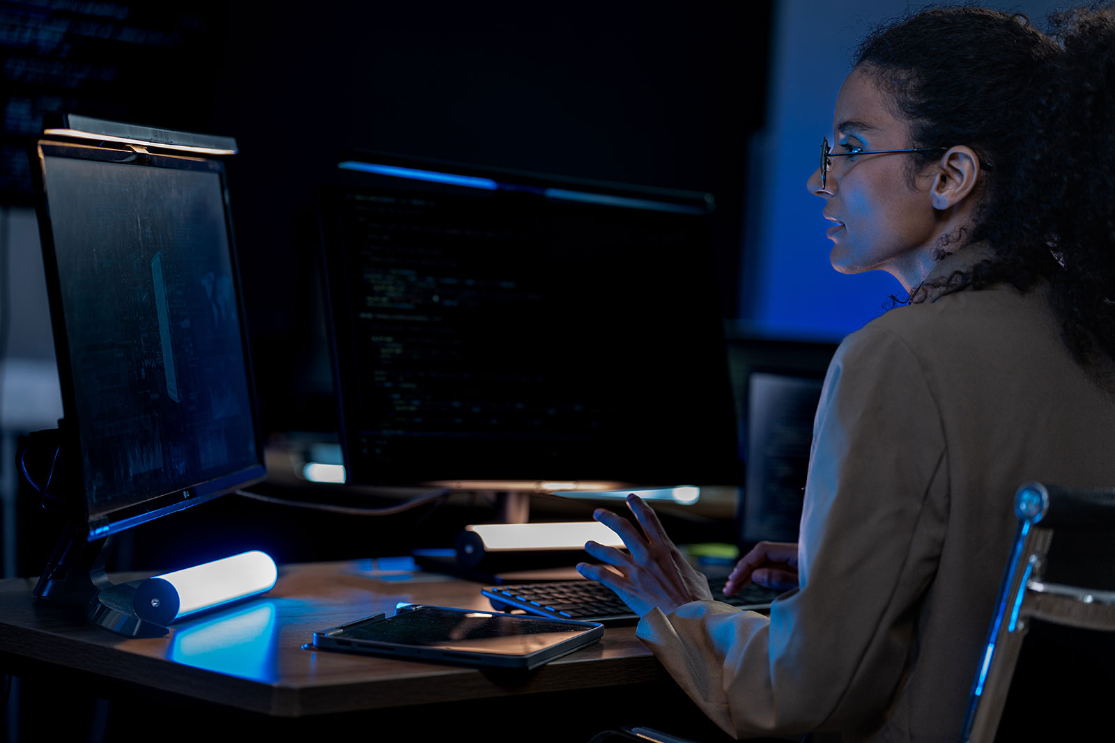 Woman in glasses typing at a keyboard, focused on multiple monitors showing code in a dimly lit office