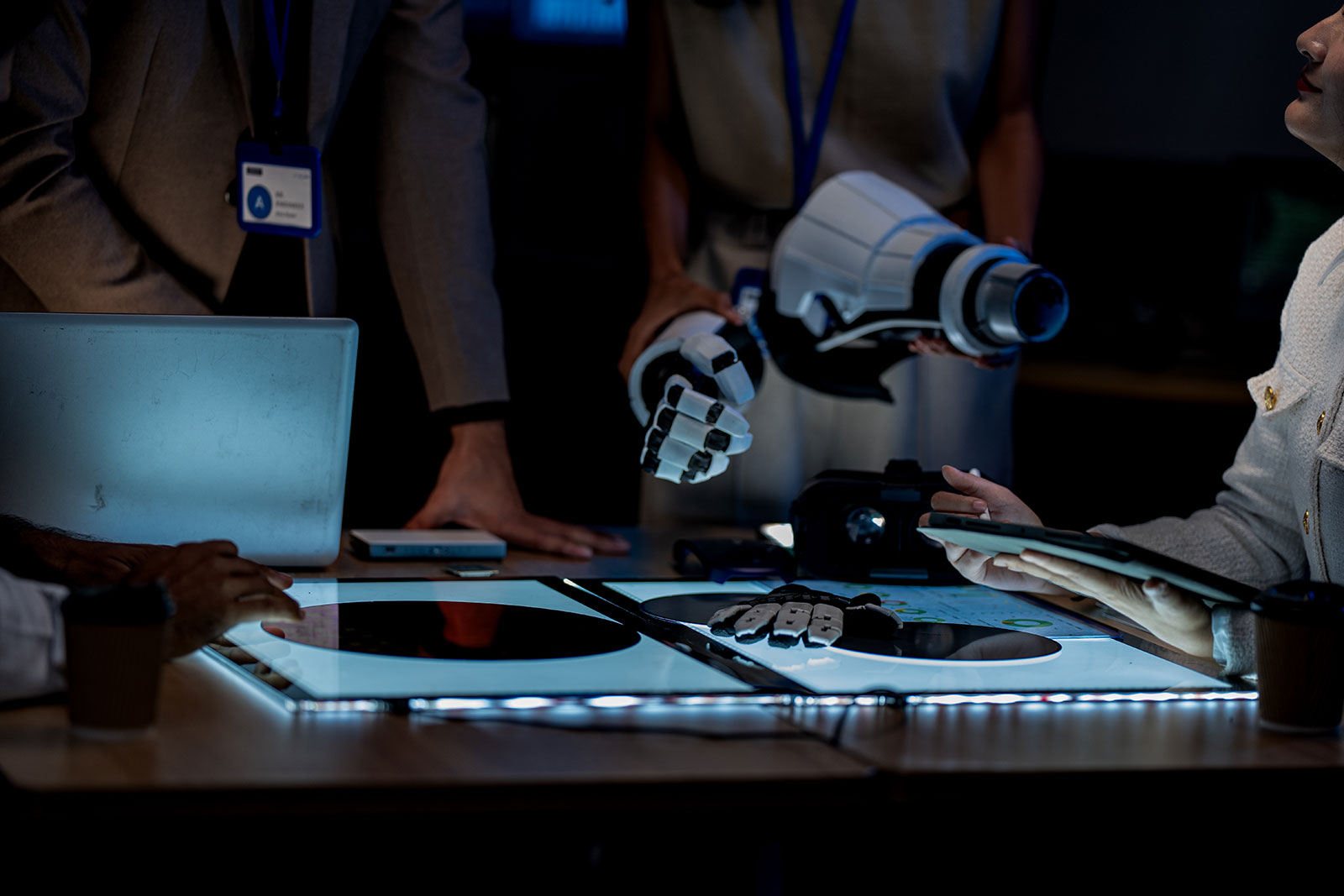 People gathered around an illuminated table with tablets and a VR headset, one holding a white robotic arm with a mechanical hand