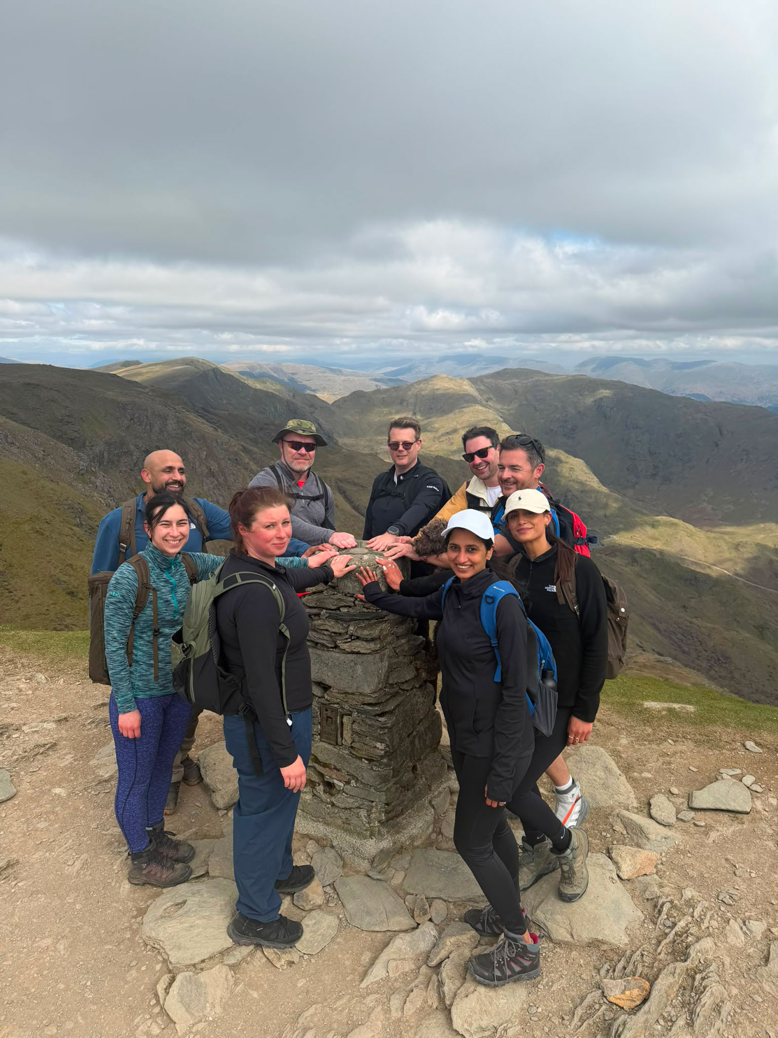 Nine members of our senior leadership team and board in walking gear, hands together on the stone trig point at the summit of the Old Man of Coniston, with Lake District fells behind