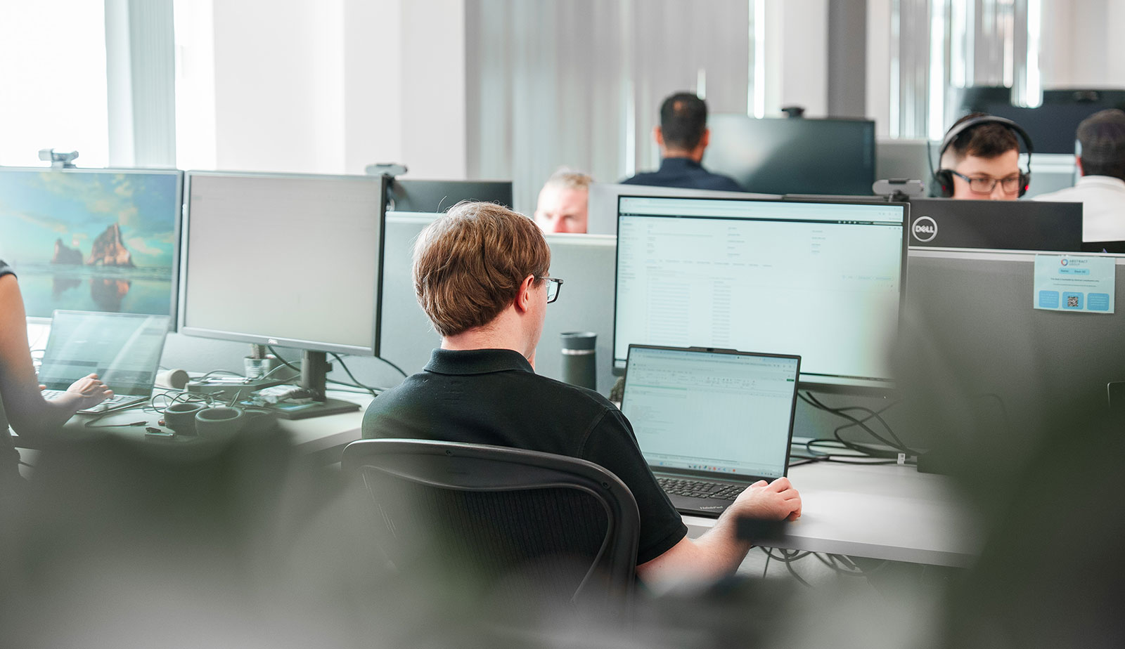 John Lamb, Automation QA Engineer, working at a laptop and monitor in our open-plan office, with colleagues at desks around him