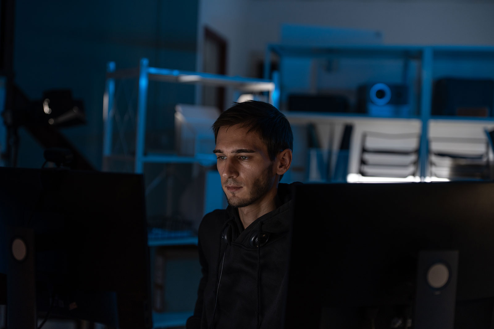 Man with headphones around his neck, concentrating on his screen between two monitors in a dimly lit office