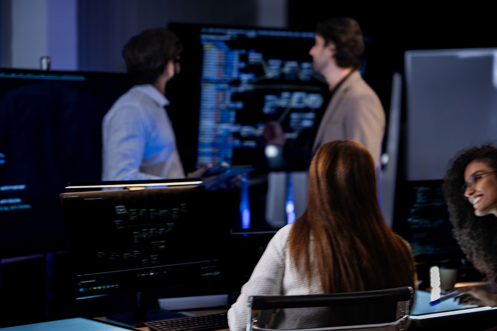 Team working in a dimly lit tech office, two colleagues discussing a large screen of code and diagrams while others work at desks
